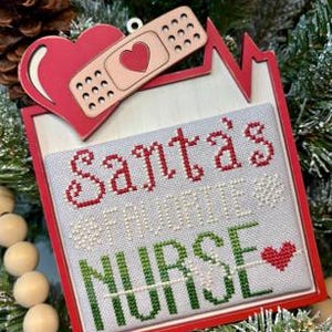 May include: A Christmas ornament with a red frame and the words "Santa's Favorite Nurse" embroidered in red and green. A red heart and bandage are attached to the top. The ornament is surrounded by pine needles and wooden beads.