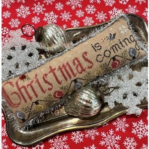 May include: A rectangular beige fabric pillow featuring the cross-stitched text "Christmas is coming" in red and black, decorated with a string of colorful light bulbs, surrounded by two silver ribbed ornaments on a metallic tray.