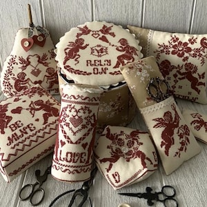 May include: A set of eight needlepoint cushions featuring a romantic theme with cherubs, hearts, and floral designs. The cushions are in shades of red and cream and have embroidered phrases like "Be Mine" and "Rely On Love".