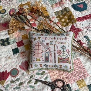 May include: A square embroidered pincushion featuring a house, flowers, and the text 'punch needle house' in brown thread. The piece is bordered by green pom-pom trim and rests on a patchwork quilt with floral patterns, accompanied by metal scissors.