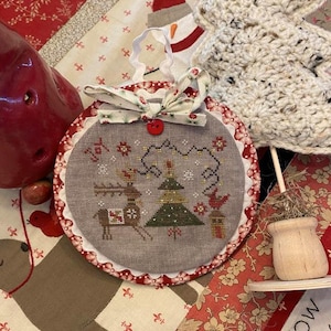 May include: A round, gray Christmas ornament with a cross-stitch design of a reindeer and Christmas tree. The ornament has a red and white scalloped edge, a fabric bow, and a red button. The background is a patchwork quilt.