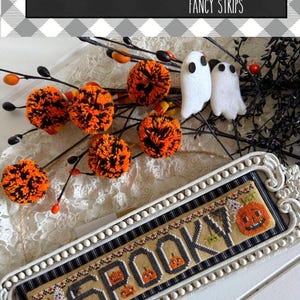 May include: A rectangular cross-stitch sampler in a white beaded frame featuring the word SPOOKY with orange jack-o'-lanterns and black spiders. The piece is displayed with orange and black yarn pom-poms and two white felt ghost figures.