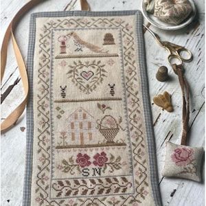 May include: A beige needlepoint project bag with a blue and white gingham border. The bag features a stitched design of a bird, a heart, a house, and flowers. The bag has a brown ribbon loop for hanging.