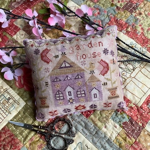 May include: A hand-stitched pillow with a light purple and brown house design and the text "Garden House". The pillow is on a patchwork quilt with pink flowers.