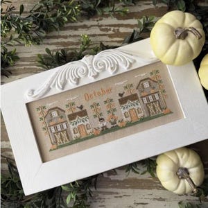 May include: A white framed cross-stitch piece with the word "October" and a village scene. The scene includes houses, pumpkins, and a scarecrow. Two small white pumpkins and greenery surround the frame.