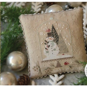 May include: A cross-stitch pillow featuring a snowman in a snow globe with the text "Let It Snow!" and "design by Brenda Gervais". The pillow is surrounded by white and silver ornaments and greenery.