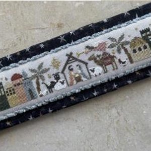 May include: A cross-stitch embroidery depicting the nativity scene with a star, a stable, a camel, sheep, and palm trees. The scene is framed with a border of stars.