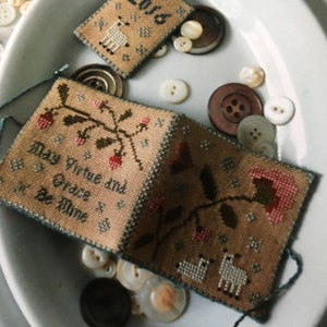 May include: A small, folded, cross-stitch sampler with a beige background and blue trim. The sampler features a floral design with a pink flower, green leaves, and red berries. The text on the sampler reads "May Virtue and Grace Be Mine".