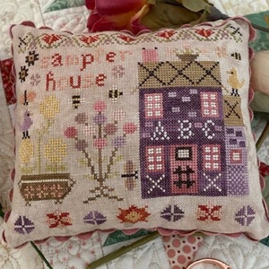 May include: A sampler cross stitch pillow with a pink, purple, and brown color scheme. The pillow features a house with a flower garden and the text "sampler house".
