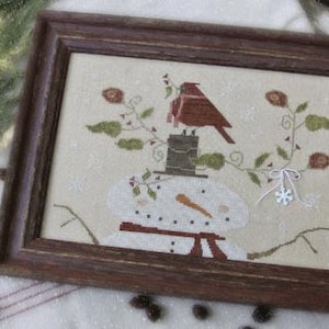 May include: A cross-stitch pattern featuring a snowman with a red cardinal perched on top of its head. The snowman is surrounded by a wintery scene with snow, branches, and berries. The pattern is titled "All Bundled Up!" and is designed by Brenda Gervais.