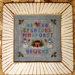 May include: A framed cross-stitch sampler with a winter theme. The sampler features the alphabet in various colors, a red bird, snowmen, a house, and holly. The frame is white with a textured design.