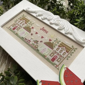 May include: A white framed cross-stitch embroidery depicting a summer scene with houses, trees, and the word "June".