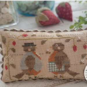 May include: A rectangular, beige cross-stitch pillow featuring two robins wearing hats and scarves, with bees and berries. The text "THE ROBINS ARE HERE!" is on the left side. The design is by Brenda Gervais.
