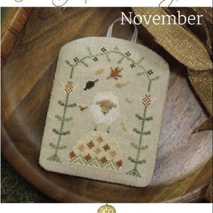 May include: A beige cross-stitch design featuring a sheep, leaves, and floral elements. The text "The Shepherd's Year November" is displayed above the design. The design is on a small, square, fabric piece with a white loop for hanging.