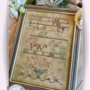 May include: A framed cross-stitch sampler titled "Clarissa's Garden Sampler" featuring a floral design with birds and the alphabet. The sampler is stitched on a beige fabric and framed in a gold-colored frame.