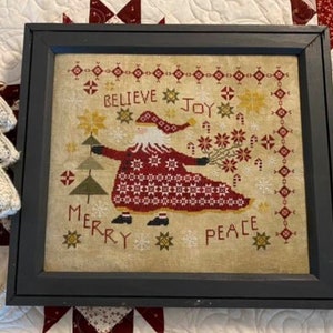 May include: A framed cross-stitch design featuring a Santa Claus figure in a red and white patterned robe, holding a sprig of holly. The design includes the words "Believe", "Joy", "Merry", and "Peace" in a vintage style font. The frame is black and the background is a cream color.