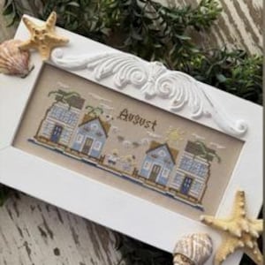 May include: A framed cross-stitch piece featuring a coastal scene with blue houses and the word "August". The white frame is adorned with decorative elements and seashells. The artwork is displayed on a wooden surface with greenery and starfish.