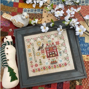 May include: A framed cross-stitch pattern with a red house, a bird, and the word "GARDEN" in a floral design. The frame is black and the background is a patchwork quilt.