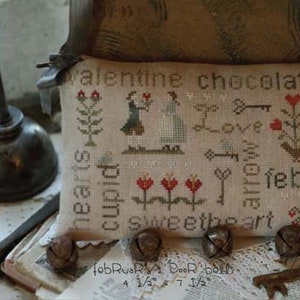 May include: A cross-stitch sampler with a beige background and colorful embroidery. The sampler features a variety of Valentine's Day themed imagery, including hearts, cupids, roses, and the words "Valentine chocolates", "Love", "Sweetheart", and "February".