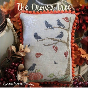 May include: A cross-stitch pillow featuring a beige background with a brown tree and black crows. The pillow has an orange trim with pom-poms. The text "The Crow's Tree" is at the top of the pillow.