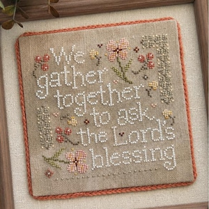 May include: A square cross-stitch embroidery on light brown linen fabric featuring the white text We gather together to ask the Lord's blessing, surrounded by small embroidered flowers in pink, yellow, and red, framed in a wooden border.