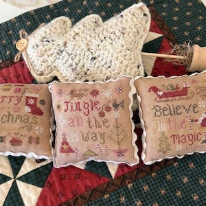 May include: Three beige Christmas-themed cross-stitch pillows with white ruffled edges. The pillows feature festive text and images, including a stocking, a sleigh, and a Christmas tree. A crocheted tree and a wooden ornament are also in the image.