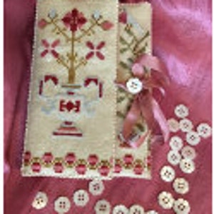 May include: A white needlepoint button box with a floral design in shades of pink, red, and gold. The box is tied closed with a pink ribbon and has a row of pearl buttons on the bottom.