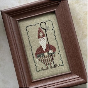 May include: A framed cross-stitch design featuring a whimsical gnome with a red hat, white beard, and striped pants holding a star-topped stick. The gnome is surrounded by a decorative border.