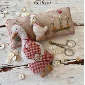May include: A handmade fabric goose with a red and white checkered apron and a smaller goose figurine rest on a beige pillow with floral embroidery. The image is from the "Animal Cracker Series" and includes the text "Design by Stacy Nash."