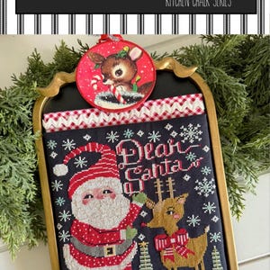 May include: A framed Christmas-themed cross-stitch featuring Santa Claus and a reindeer, with the words "Dear Santa." The design includes snowflakes and Christmas trees. A round ornament with a reindeer is at the top.