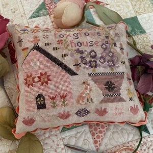 May include: A hand-stitched pillow with a pink house, a fox, and a pot of purple flowers. The pillow is decorated with a scalloped edge and the text "Foxglove House".
