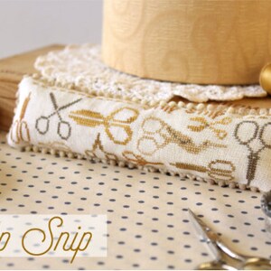 May include: A white needlepoint pillow with a brown and gold embroidery design of scissors and other sewing tools. The pillow is on a white and blue polka dot fabric with a wooden box and a spool of thread in the background. The text "OH1255 Snip Snip" is printed below the pillow.