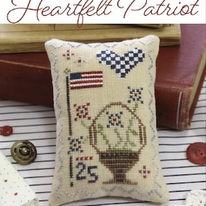 May include: A beige pillow with a cross-stitch design featuring a heart, a flag, flowers, and a basket. The pillow is on a striped surface with books and other craft supplies.