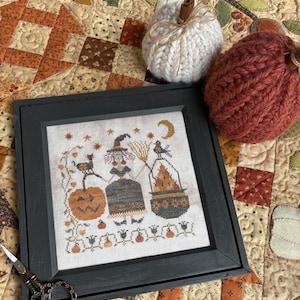 May include: A framed cross-stitch featuring a Halloween theme with a witch, black cat, and pumpkin. The frame is dark gray. Two knitted pumpkins and a white knitted pumpkin with a cinnamon stick are also in the image. A pair of scissors is in the lower left corner.