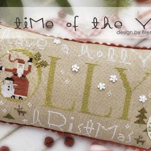 May include: A cross-stitch pattern on a beige pillow with a red border. The pattern features a Santa Claus character holding a candy cane and a snowman. The text "Holly" is stitched in green and the text "Merry Christmas" is stitched in white.