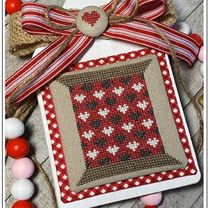 May include: A white wooden sign with a red and white gingham border. A small square piece of fabric with a pattern of red, white, and black hearts is attached to the center of the sign. A red and white striped ribbon with a burlap bow and a red heart button is attached to the top of the sign. "Original Design by Crafty Bluebonnet Designs" is printed at the bottom of the sign.