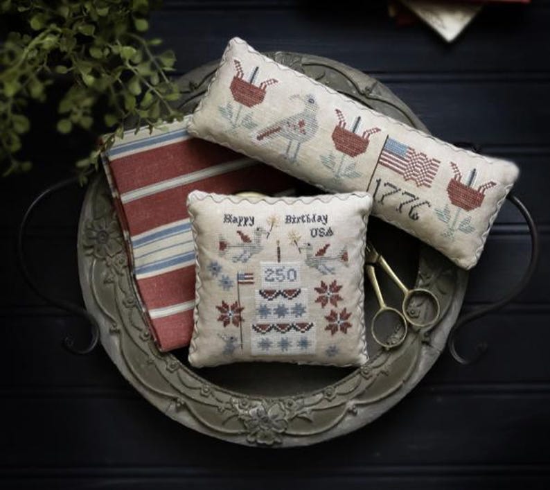 May include: A decorative tray holds three embroidered pillows and a striped fabric. The pillows feature patriotic designs with birds, flags, and the text "Happy Birthday USA 250" and "1776". A pair of scissors rests on the tray.