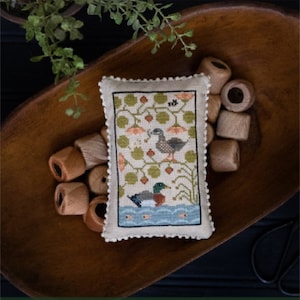 May include: A cross-stitch sampler with a wooden bowl and spools of thread. The sampler features two ducks in a pond with green foliage and flowers. The text "Quack Stack" is below the sampler. The text "Original Designs by Paulette Stewart" is below the text "Quack Stack".