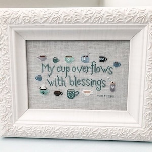 May include: White framed cross-stitch embroidery with the text "My cup overflows with blessings" and the scripture reference "Psalm 23:5". The embroidery features a variety of blue and green cups and mugs.