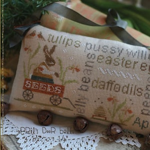 May include: A beige decorative pillow with cross-stitch embroidery featuring a bunny pulling a wagon labeled "SEEDS." The pillow includes the words "tulips," "pussy willow," "Easter eggs," and "April."