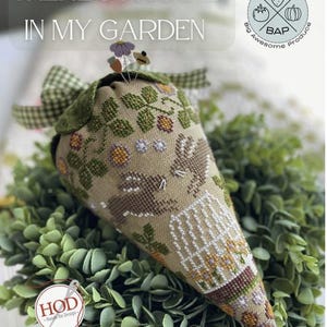 May include: A cross-stitch pattern of a cone-shaped pincushion with a hare and flowers. The pincushion is surrounded by green foliage. The text "There's a Hare in My Garden" is at the top of the image. The text "Big Awesome Produce" and "BAP" are at the top right of the image. The text "HOD - Hang On Design" is at the bottom left of the image.
