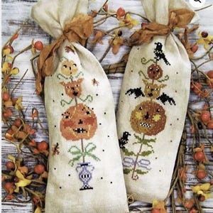 May include: Two beige fabric bags with stitched Halloween pumpkin designs. The pumpkins are orange with black details, and the bags are tied with brown ribbon. The background includes fall foliage with orange berries.