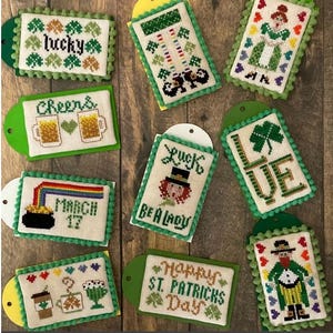 May include: A collection of St. Patrick's Day themed cross-stitch tags. Each tag features a unique design, including shamrocks, leprechauns, rainbows, and text like "Lucky," "Cheers," and "Happy St. Patrick's Day." The tags have green borders and are attached to green or yellow cardstock.