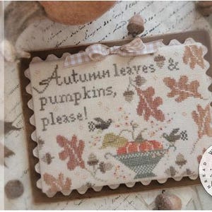 May include: A rectangular cross-stitch sampler on cream fabric with scalloped edges, featuring the text Autumn leaves & pumpkins, please! in dark brown, surrounded by embroidered orange pumpkins, brown oak leaves, acorns, and two small black birds.