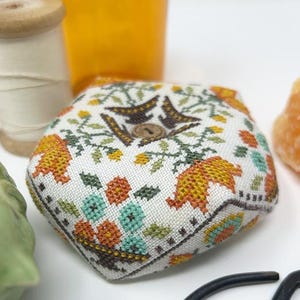 May include: A pentagonal pincushion featuring a white fabric base with cross-stitched floral patterns in orange, yellow, green, and teal. The center of the pincushion is adorned with a small, round, brown wooden button, adding a textured detail.