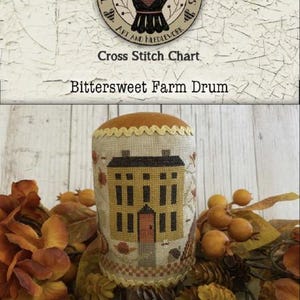 May include: A cross stitch chart titled "Bittersweet Farm Drum" featuring a detailed illustration of a yellow house with a red door. The chart is surrounded by fall foliage and berries. The image includes the text "Teresa Kogut's Creative Whims".