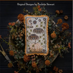 May include: A cross-stitch sampler with a cream background and orange fringe. The design features three brown spiders with black eyes, a crescent moon, and a pumpkin. The text "Jump Clump" is below the sampler.