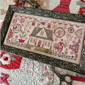 May include: Framed cross-stitch sampler featuring a detailed house, flowers, and a bird. The artwork is in a dark green ornate frame. The color palette includes shades of pink, brown, and green, creating a vintage aesthetic. The sampler is displayed on a quilted background.