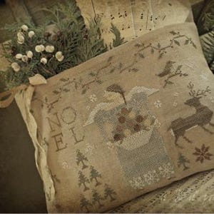 May include: A beige pillow with a cross-stitch design featuring an angel holding a basket of ornaments, a deer, and the word "Noel". The pillow is surrounded by greenery and a piece of sheet music.