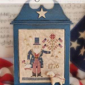 May include: A framed cross-stitch design with a patriotic theme. The blue frame holds a stitched image of Uncle Sam, flags, and the year 1776. A small sheep figurine is in front of the frame. The text "One Flag, One Land!" is at the top.
